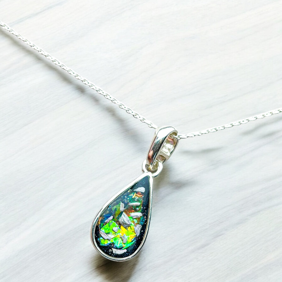 A silver teardrop pendant necklace featuring a clear resin inlay containing brown hair and tiny iridescent flecks, hanging from a silver chain against a soft gray stone background. Handmade in Wales customisable with breastmilk, cremation ashes, umbilical cord, placenta, teeth, hair/fur