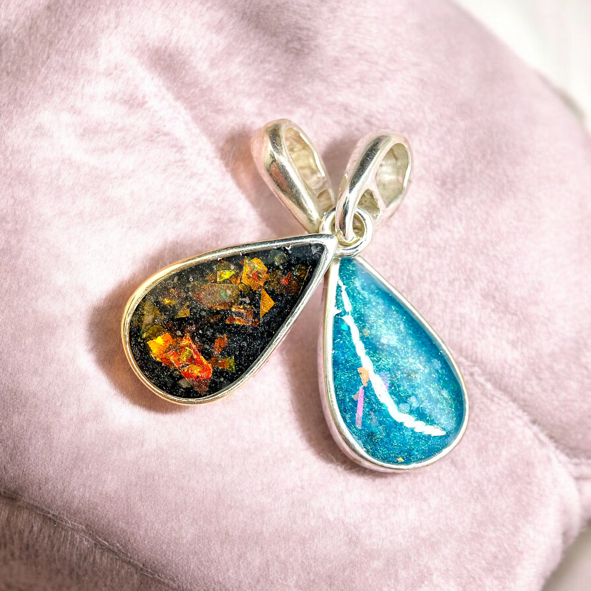 A silver teardrop pendant necklace featuring a clear resin inlay containing brown hair and tiny iridescent flecks, hanging from a silver chain against a soft gray stone background. Handmade in Wales customisable with breastmilk, cremation ashes, umbilical cord, placenta, teeth, hair/fur