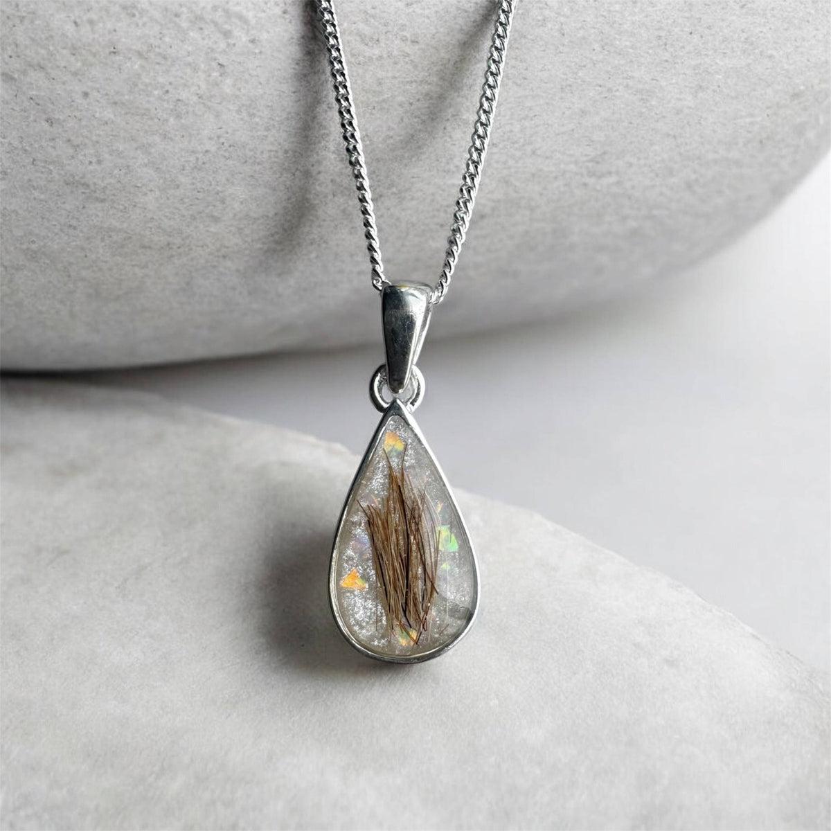 A silver teardrop pendant necklace featuring a clear resin inlay containing brown hair and tiny iridescent flecks, hanging from a silver chain against a soft gray stone background. Handmade in Wales customisable with breastmilk, cremation ashes, umbilical cord, placenta, teeth, hair/fur