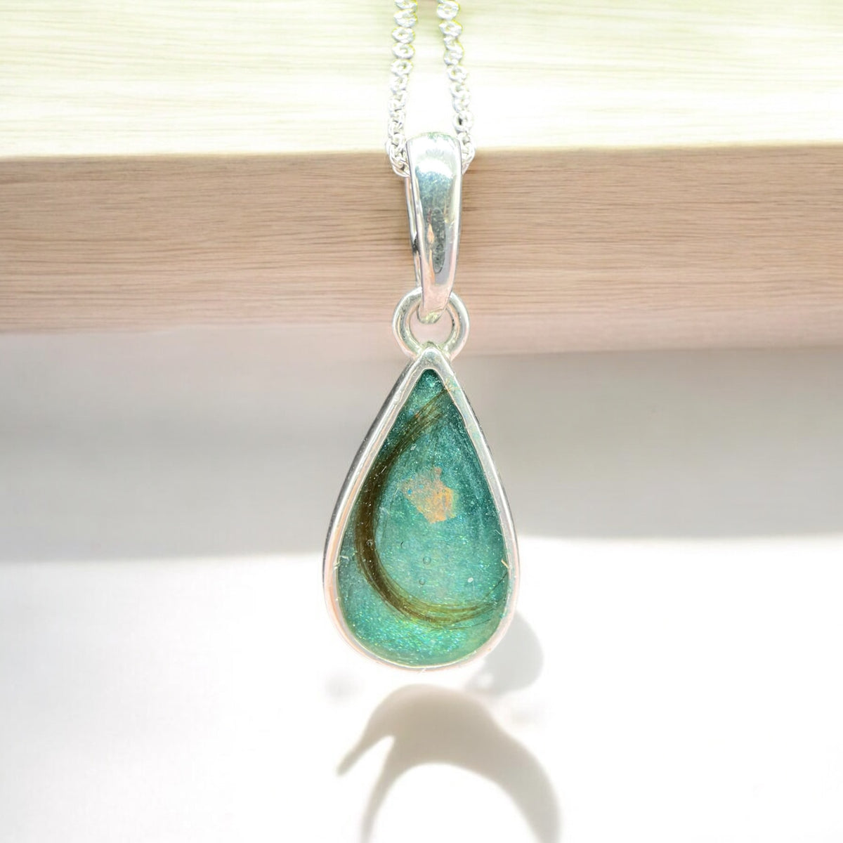 A silver teardrop pendant necklace featuring a clear resin inlay containing brown hair and tiny iridescent flecks, hanging from a silver chain against a soft gray stone background. Handmade in Wales customisable with breastmilk, cremation ashes, umbilical cord, placenta, teeth, hair/fur