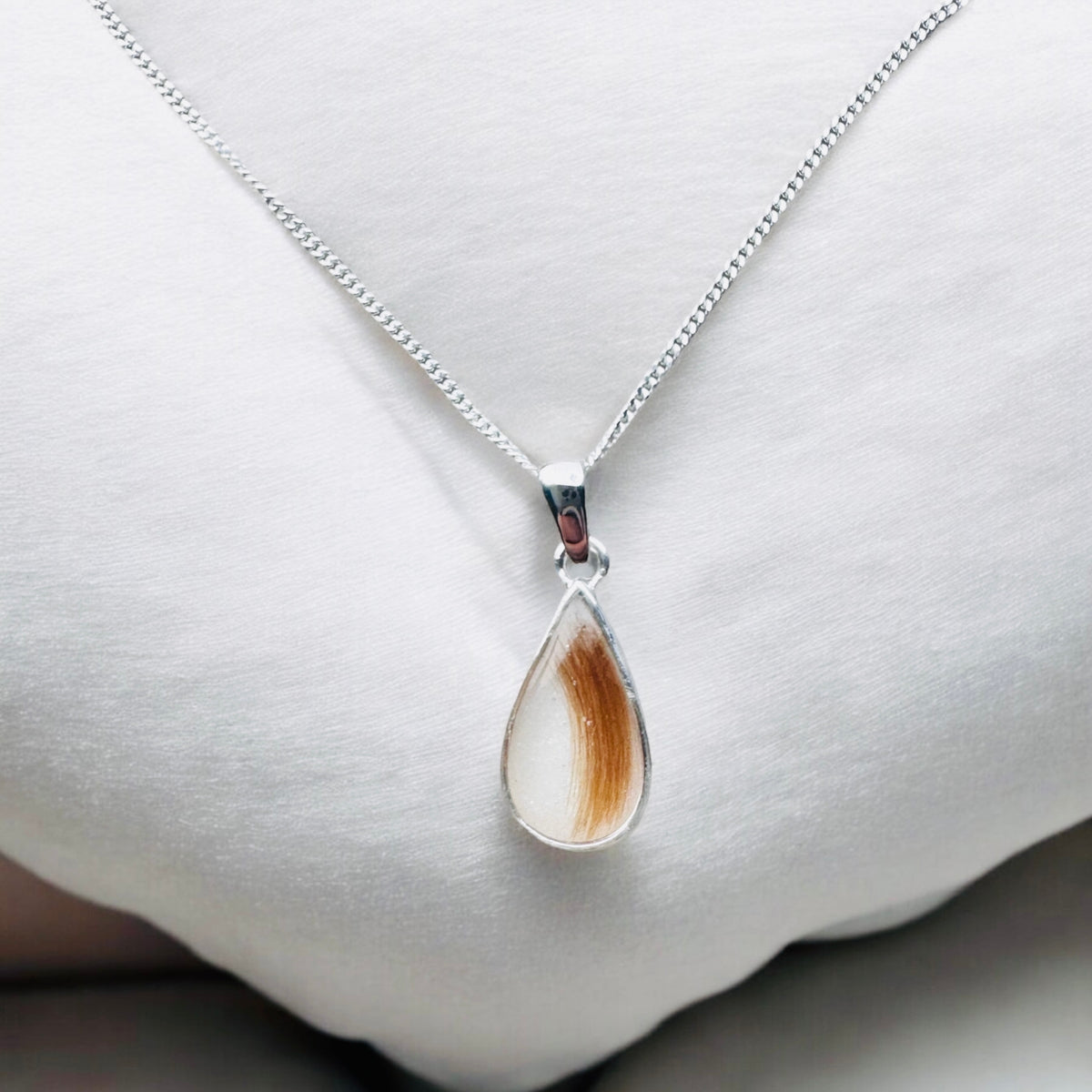 A silver teardrop pendant necklace featuring a clear resin inlay containing brown hair and tiny iridescent flecks, hanging from a silver chain against a soft gray stone background. Handmade in Wales customisable with breastmilk, cremation ashes, umbilical cord, placenta, teeth, hair/fur